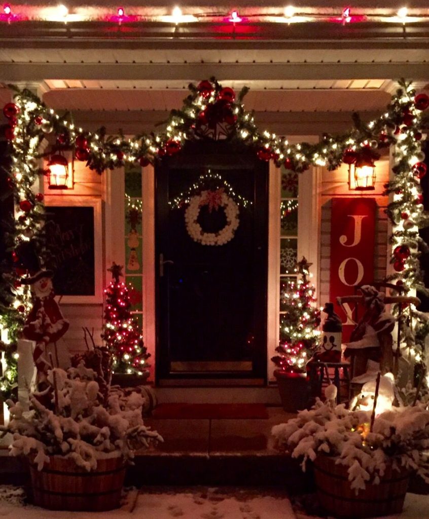 farmhouse style Christmas porch with a vintage sled or sleigh, wooden sign, lanterns with candles, evergreen boughs, and pinecones